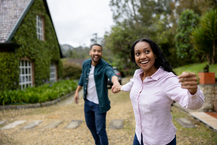 Happy young wife happily pulls her husband's arm towards a home they can afford after calculating how much mortgage they can afford.