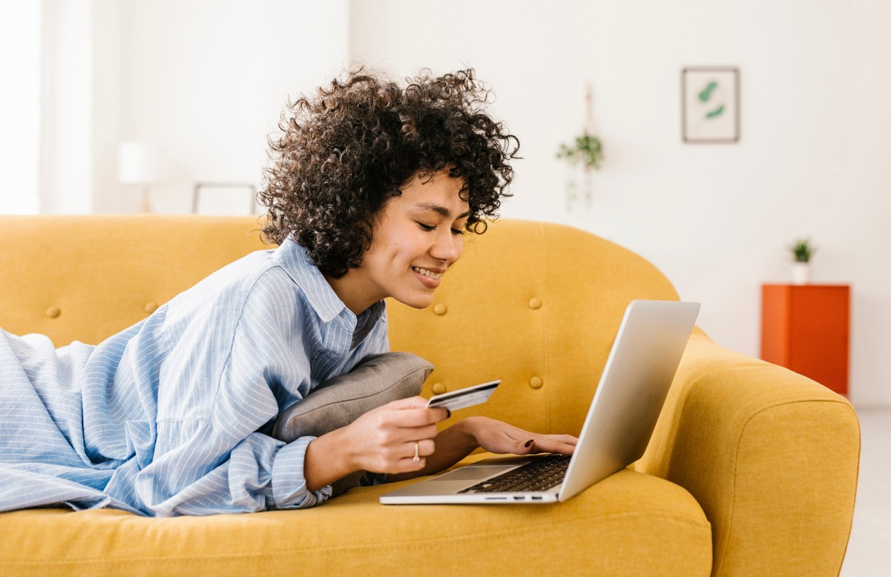 A woman lays down on a yellow couch with her laptop in front of her while holding a credit card