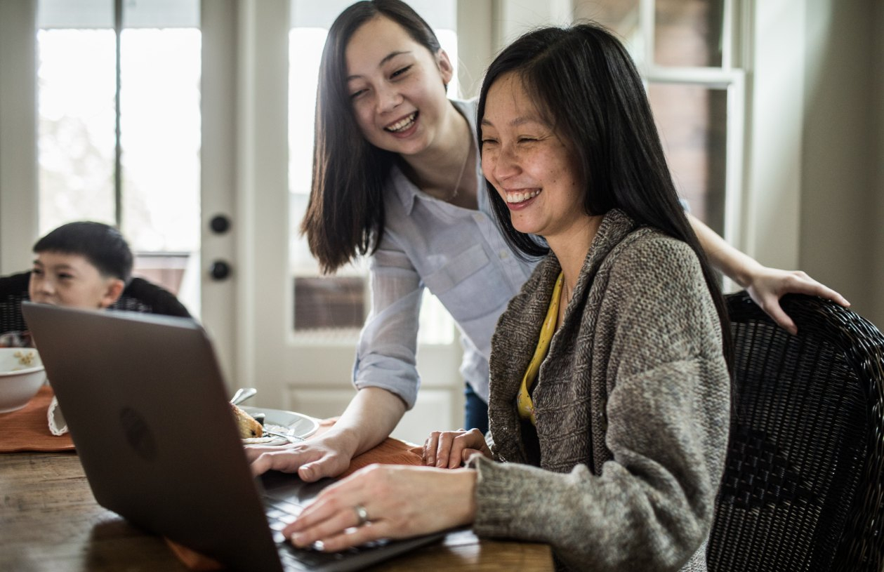 A mother sits at a table on her laptop while her daughter looks over her shoulder smiling