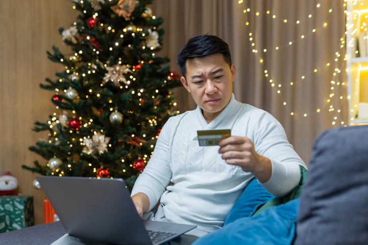 During holiday season, man looks worried looking at his credit card and online accounts on his laptop.