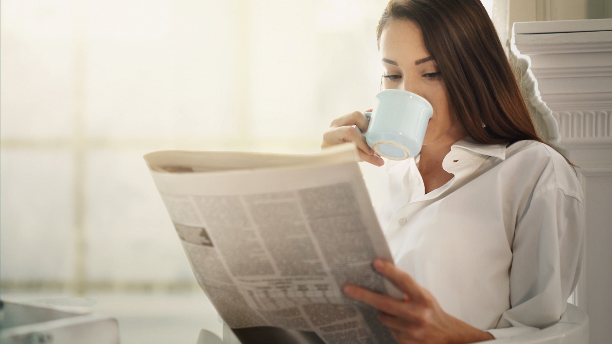 A woman sits down in her home while drinking her morning coffee and reading the newspaper
