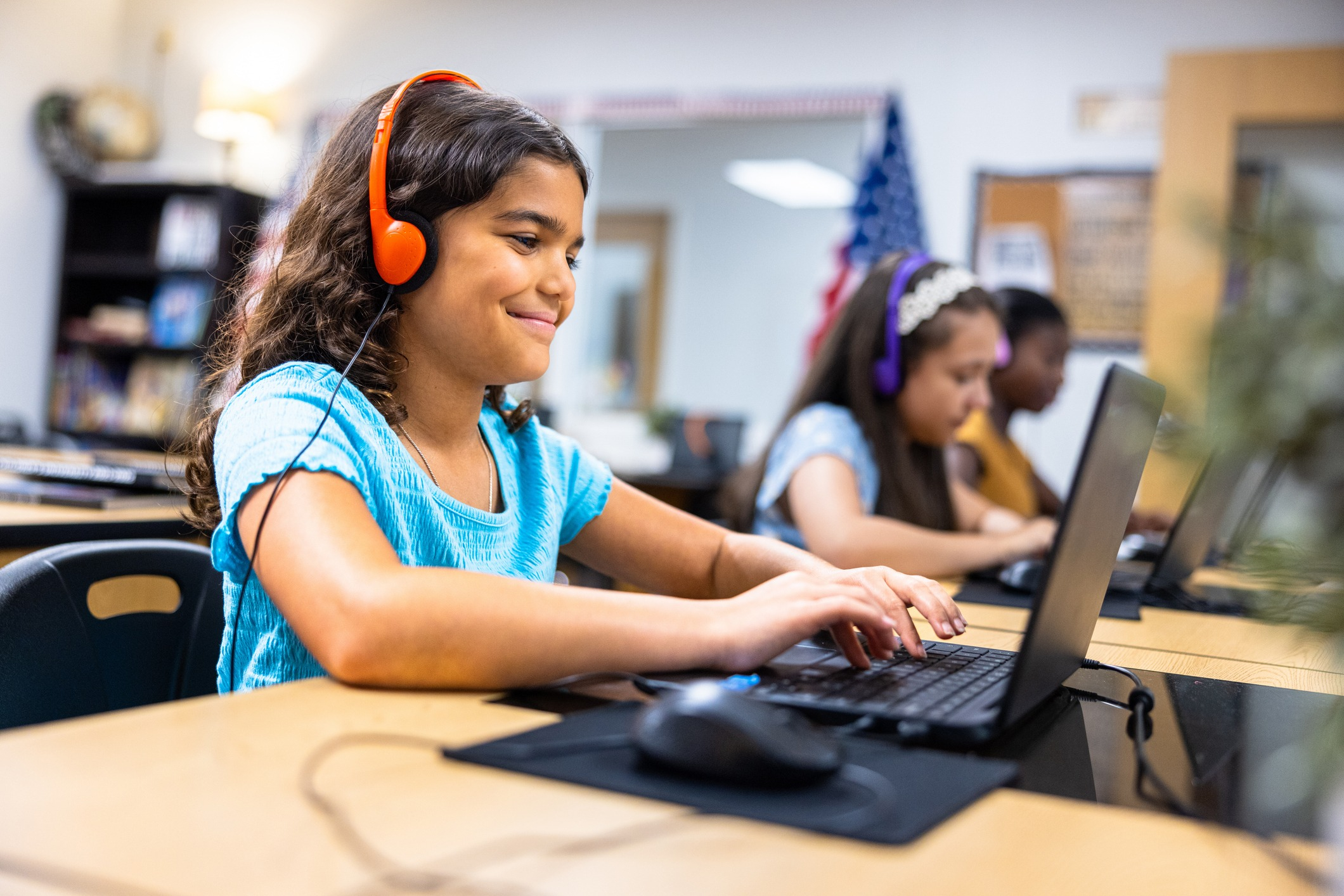 Female elementary school student happily works on  homework at school on a laptop with headphones in. 