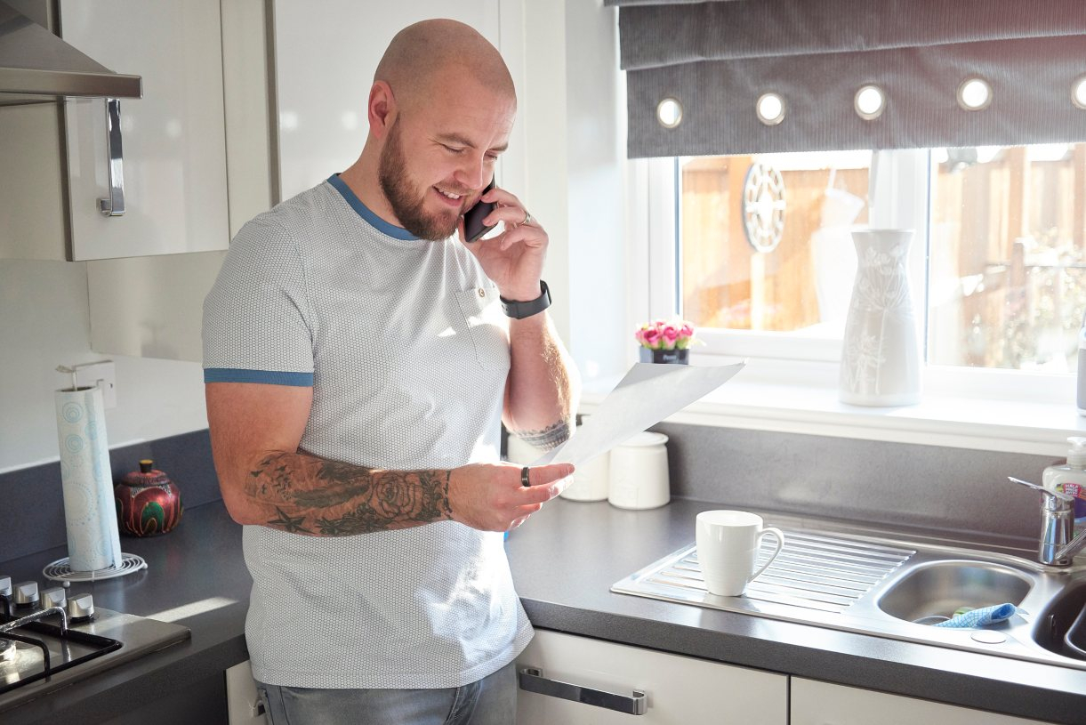 A man stand his in kitchen holding personal loan information and speaking on the phone to refinance his loan