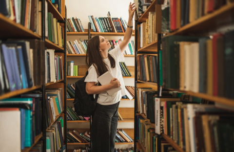 A student stands in between two bookshelves in a library, reaching up to grab a book on the top shelf on the right
