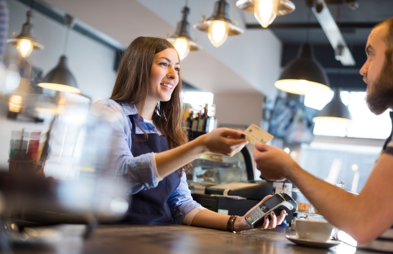 A woman working at a coffee shop takes a debit card as payment from a man ordering.