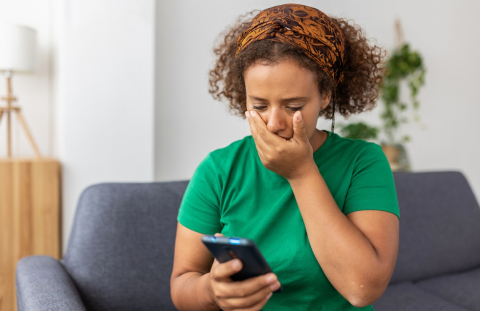A woman sitting on her couch at home looks distraught while looking at her phone