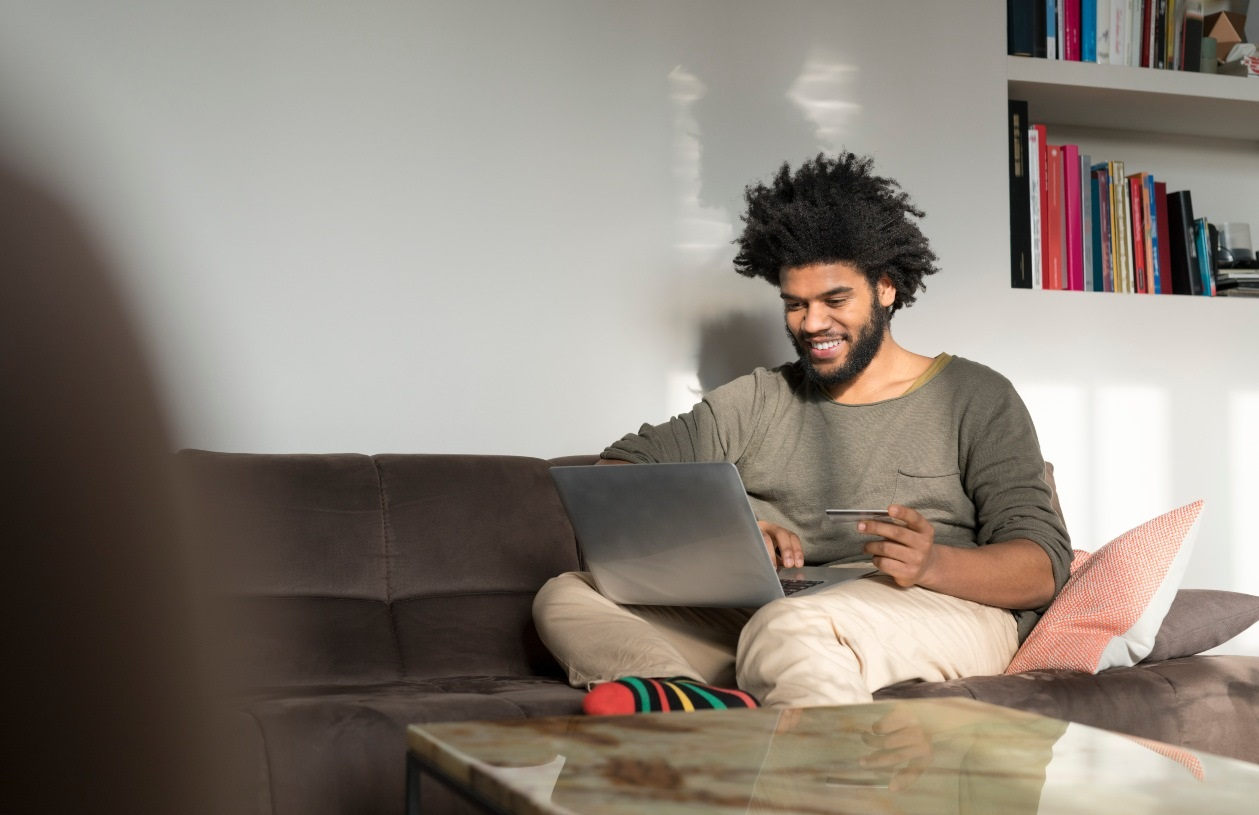 Young man sits on couch in living room. He smiles as he holds his credit card and looks at his laptop resting on his lap.
