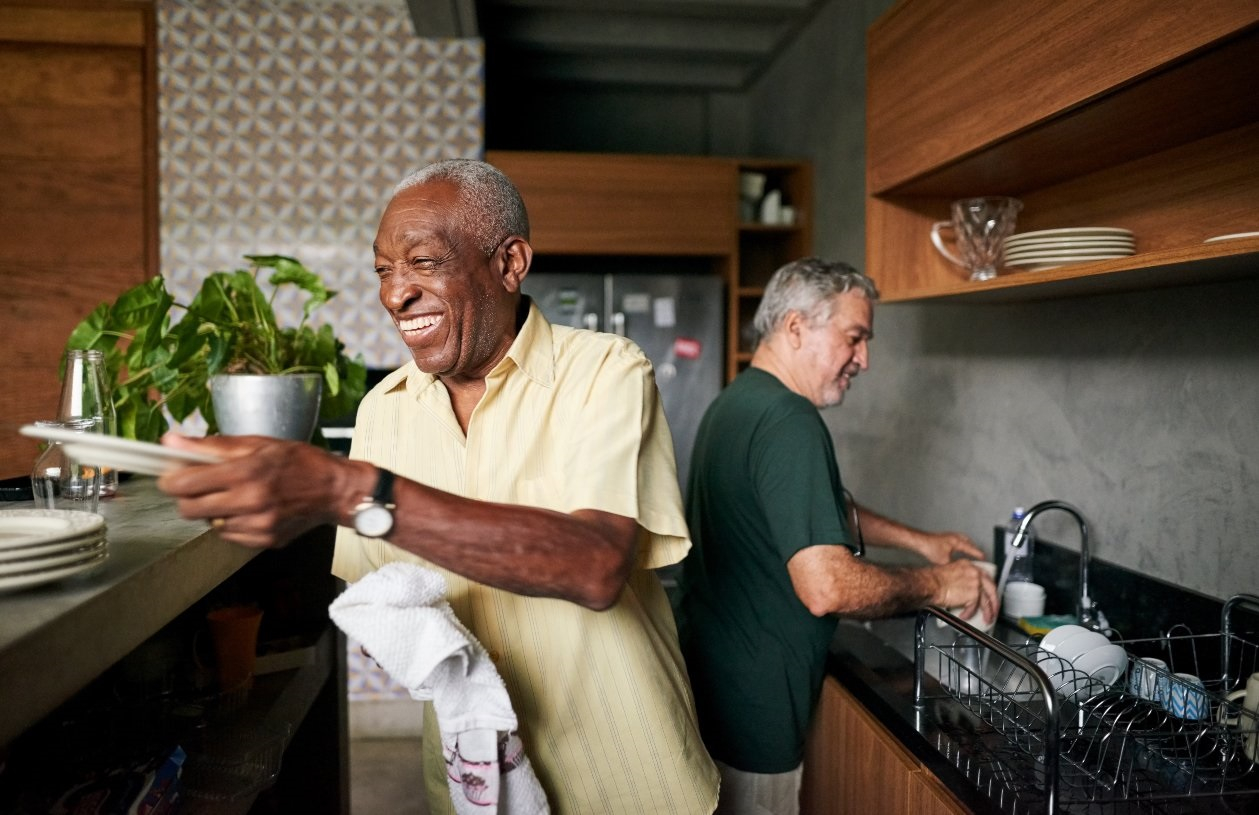 Mature couple washes and dries the dishes in their kitchen together. One man dries plates as he smiles, while the other man talks as he hand washes a cup over the sink. 