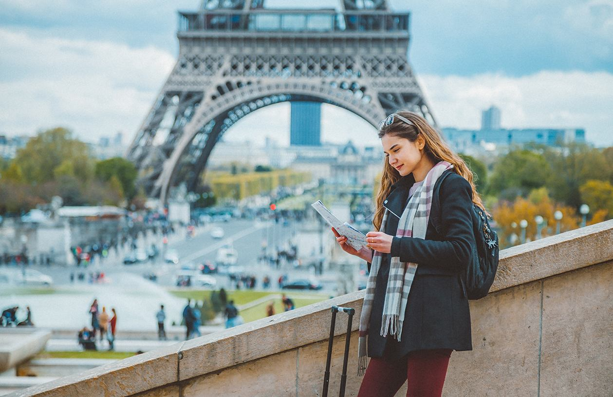 young woman stands on a bridge with the Eiffel Tower in the background looking at map next to her suitcase 