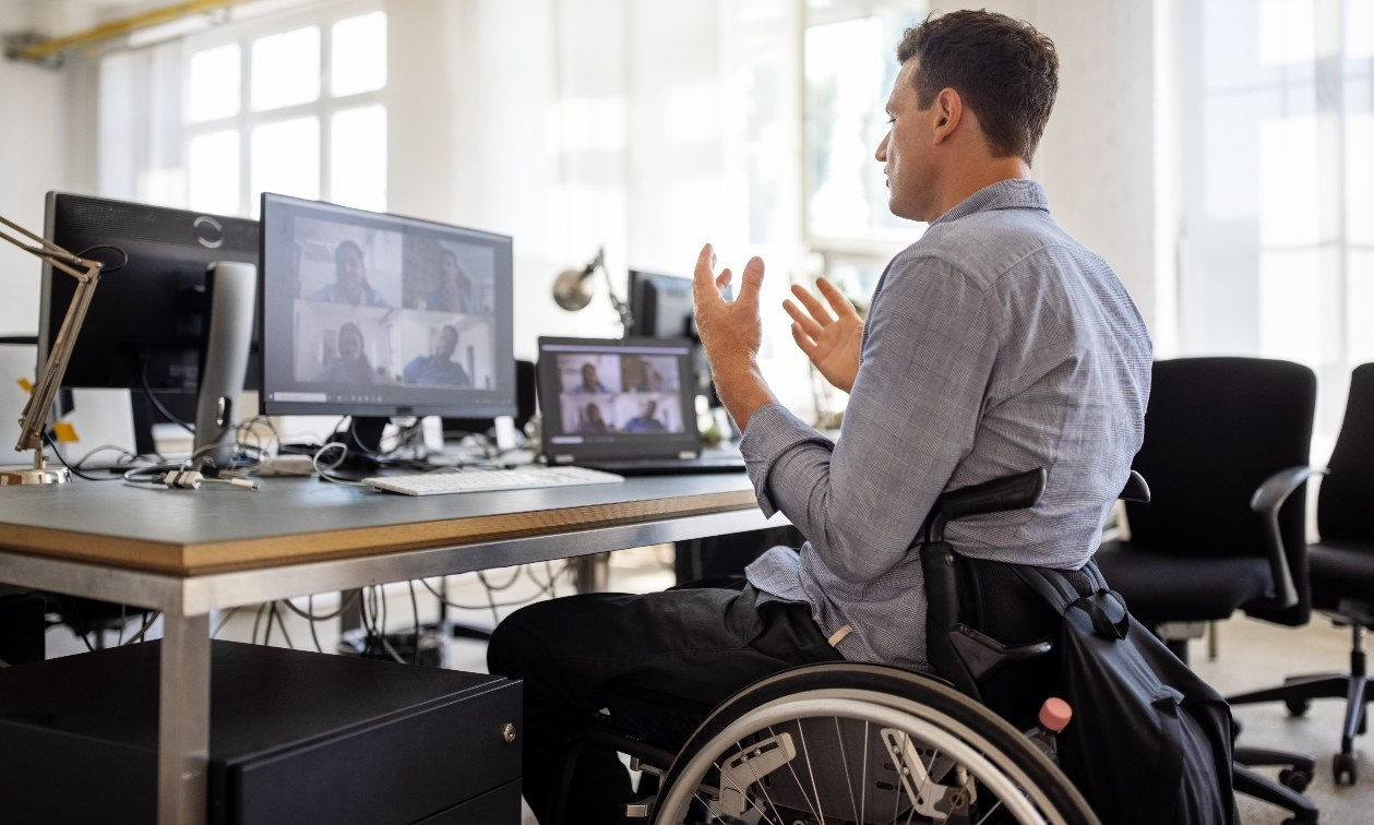 Young adult business man sits in his wheelchair at his work desk. He gestures to his computer screen where his teammates are on a virtual call. 