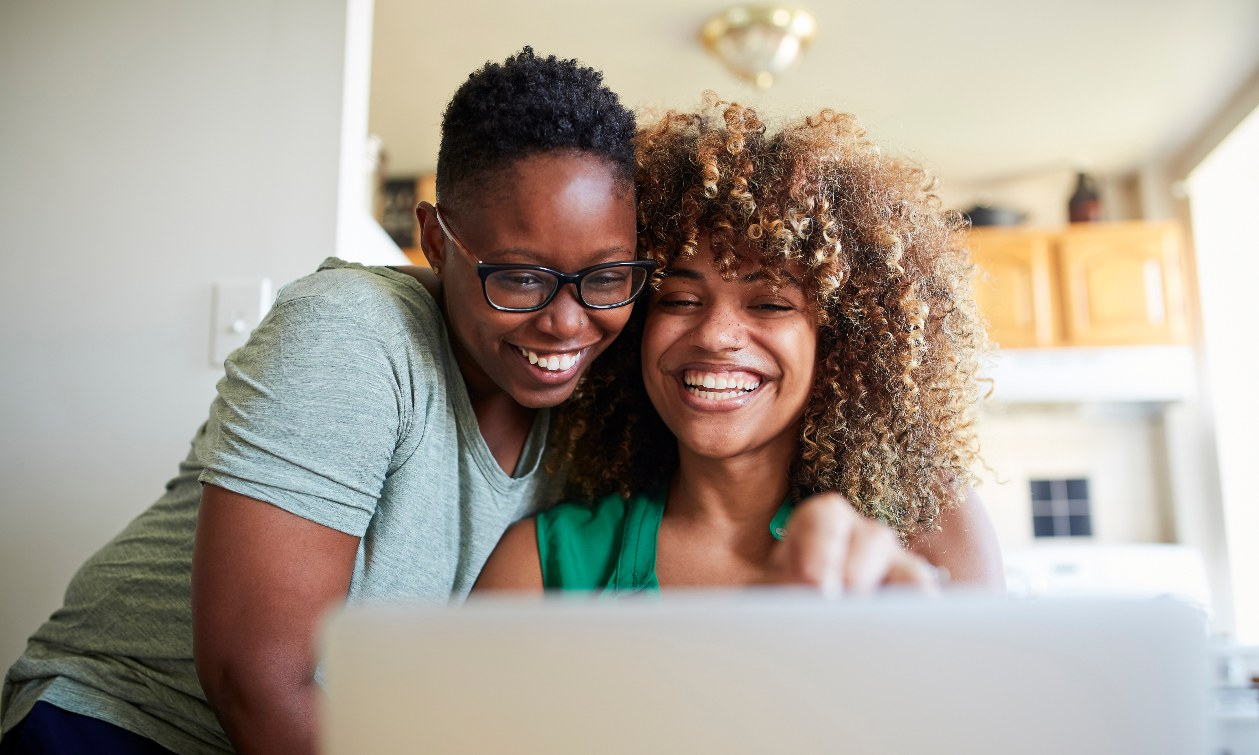 Two women sit closely as they smile at a shared laptop screen. 