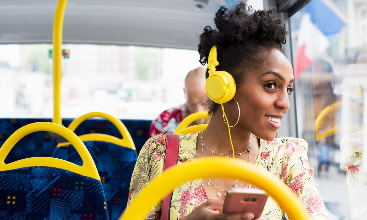 Young woman looks out the window of a bus with yellow earphones on her head. She holds her cellphone in her hand and smiles. 