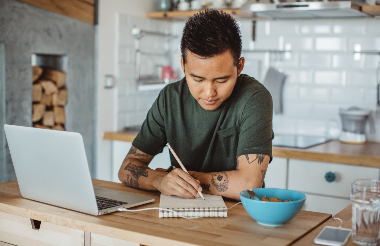 Young man sits at kitchen counter with laptop before him. He writes a list on his paper pad with a pencil. 