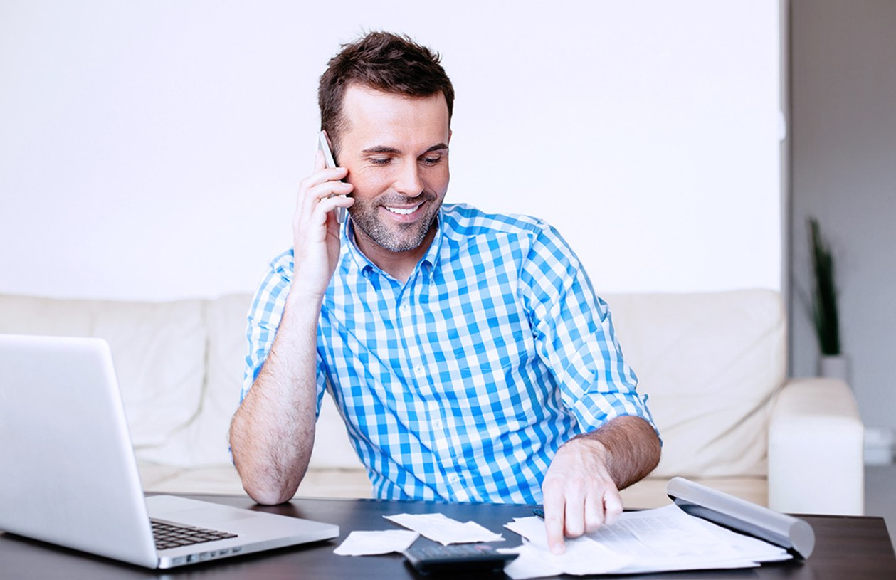 Smiling man in blue and white checkered shirt on mobile phone paying bills on his laptop
