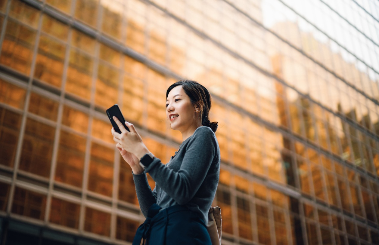A woman stands outside while looking down at her phone and smiling