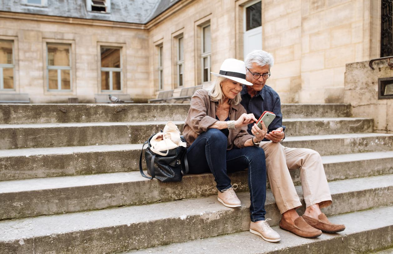 Mature husband and wife sit on steps in Paris, taking a selfie.  
