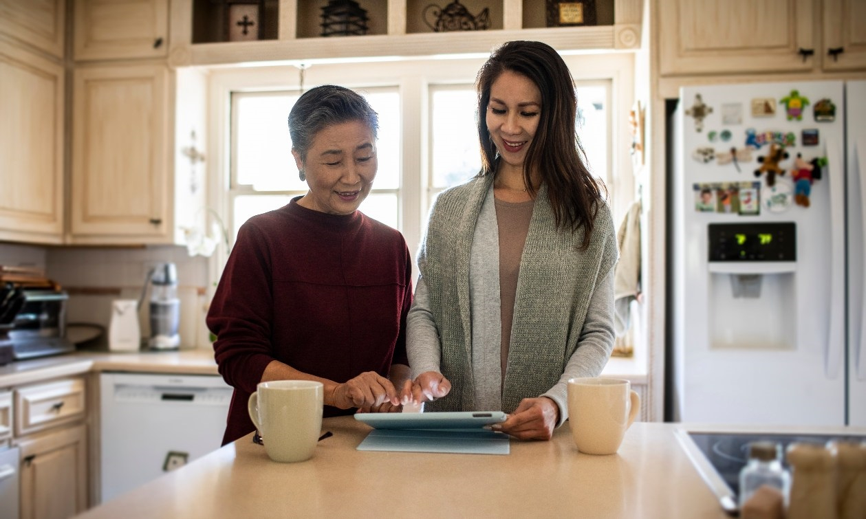Woman stands at the kitchen counter holding an iPad. She shows her mom how to use the device as they sip on coffee together. 