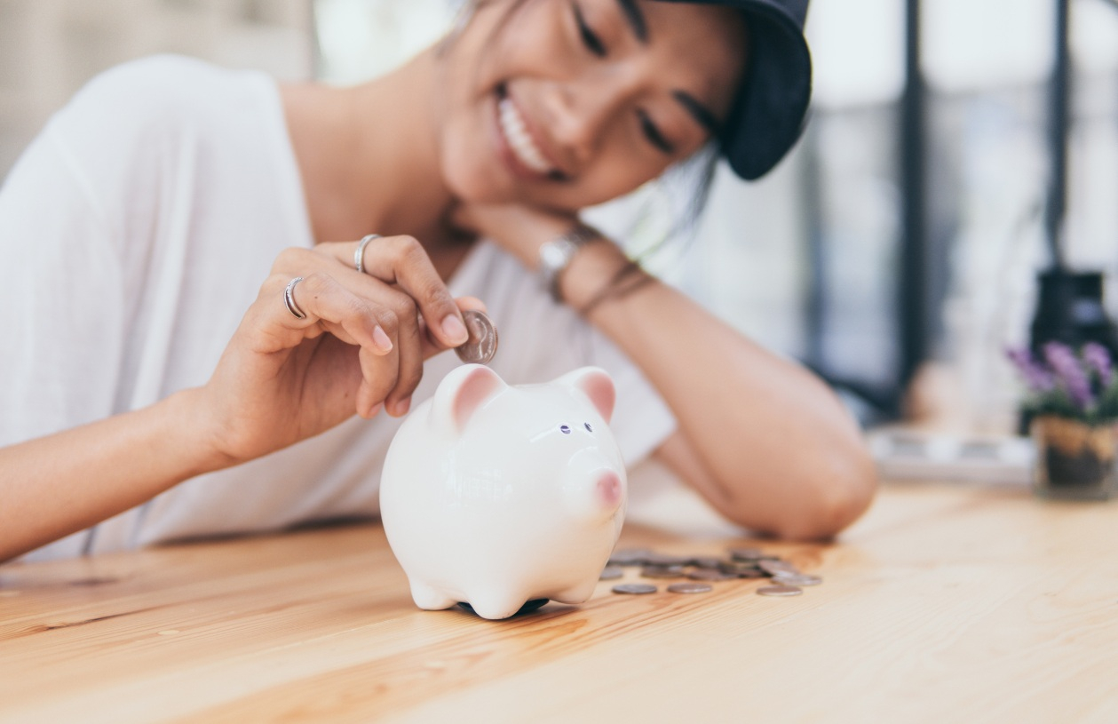 A woman smiles while putting a coin into a piggy bank on a table.