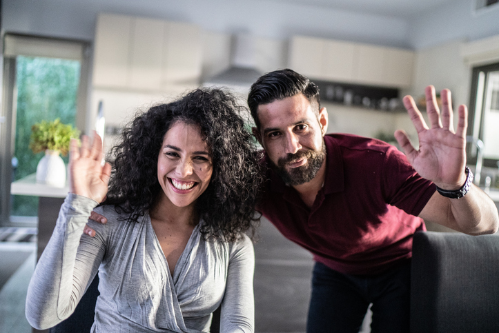 Young smiling couple both hold up five fingers in their home, reinforcing the five benefits of choosing a credit union home loan.