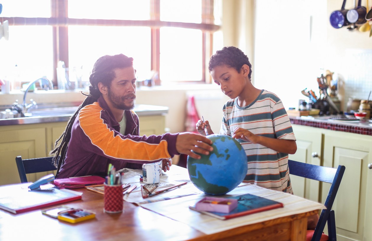 A father sits at the kitchen table with his young son, as they paint a globe together for a school project. School books and paint surround them. 