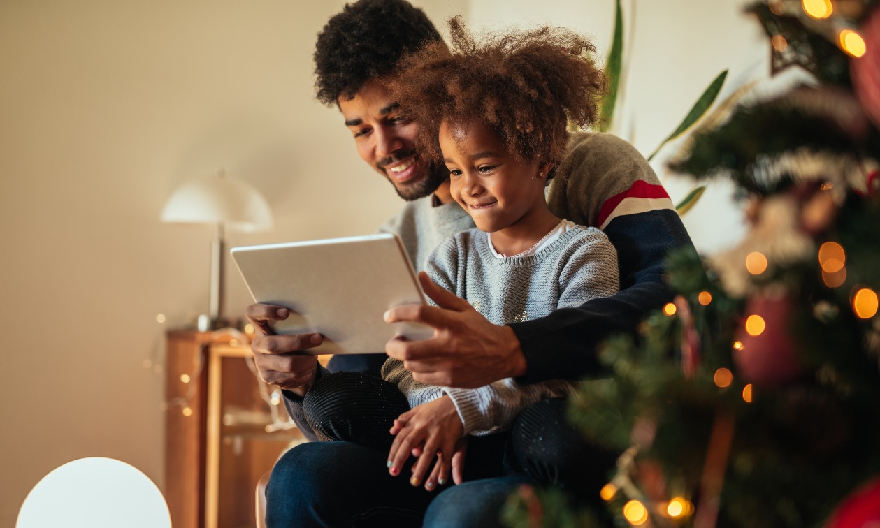 A young man sits with a little girl next to a Christmas tree. They both look at a tablet, smiling.
