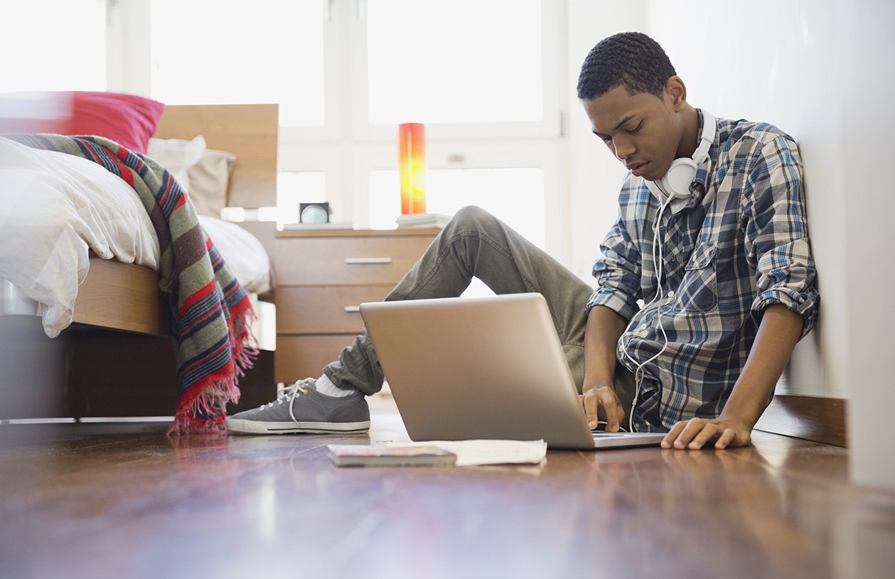 teenage boy sitting on the floor of his bedroom preparing college finances on his laptop