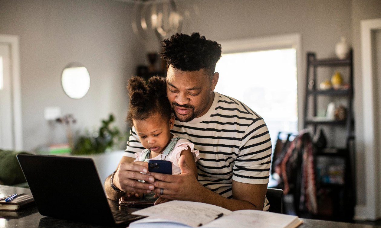 A father sits at the table with his baby in his lap. He holds his cell phone in his hands, and the baby looks at the screen before him. Papers, books, and a laptop are strewn across the table in front of them. 