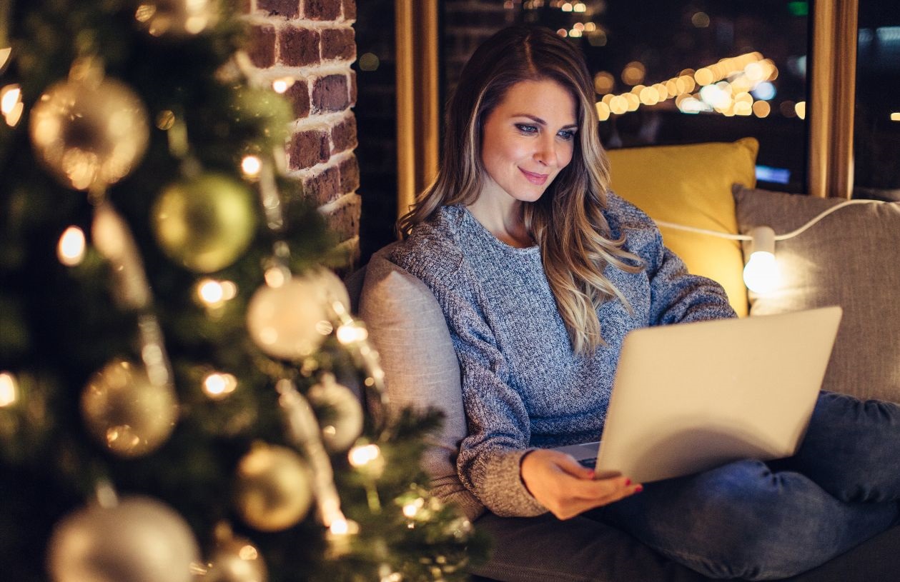 Young woman sits on her couch at night, looking at her laptop smiling. Her living room is lit with holiday lights and a festive holiday tree. 