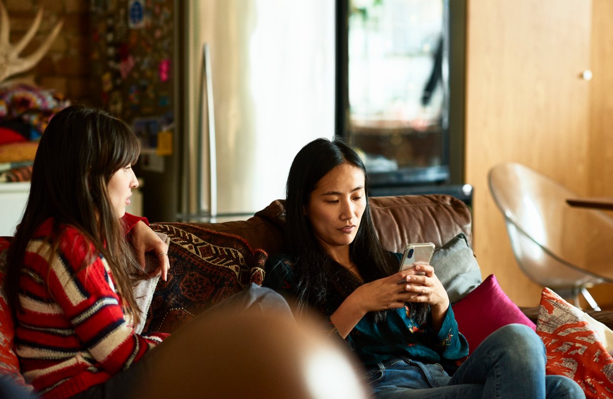 two women sit on couch while one uses her smartphone